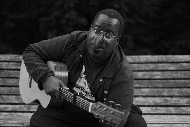 Daudi Matsiko sat on a bench with a guitar in his arms, with a beak for a nose.