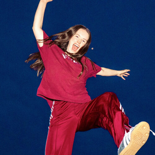 Sigrid wearing a matching t-shirt and sweatpants jumping up with one leg bent and her hands in the air.