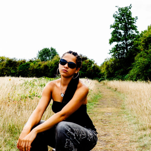 Jaya wearing sunglasses, jeans and one-shoulder top sat on a stool in a field.