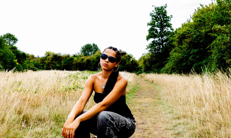Jaya wearing sunglasses, jeans and one-shoulder top sat on a stool in a field.