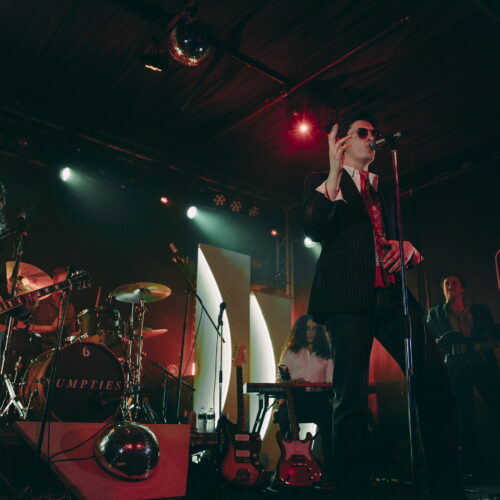 A band of five males on stage - a singer, guitarist, bassist, keyboardist, and drummer.