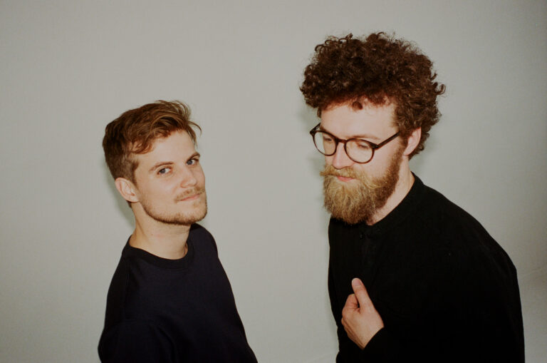 An image of two men dressed in black, the man on the left is looking into the camera while the one on the right has his hand on his chest.