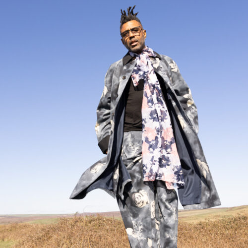 Omar standing in an open field wearing a patterned gray-blue coat and floral scarf standing in open field with dry grass and hills