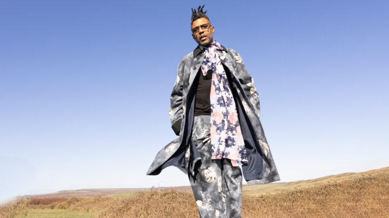 Omar standing in an open field wearing a patterned gray-blue coat and floral scarf standing in open field with dry grass and hills