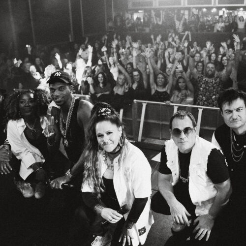 A black and white photo of six performers posing in front of a concert stage, facing the camera.