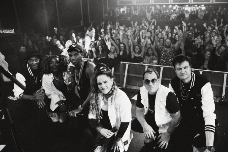 A black and white photo of six performers posing in front of a concert stage, facing the camera.