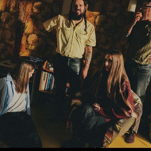 Four people in a retro-style living room with floral wallpaper and patterned furniture. Two are seated, and two are standing near a small bookshelf and side table.
