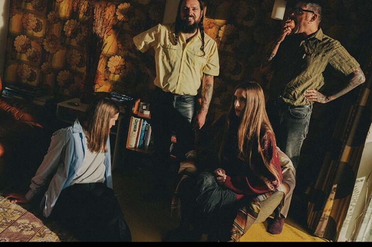 Four people in a retro-style living room with floral wallpaper and patterned furniture. Two are seated, and two are standing near a small bookshelf and side table.