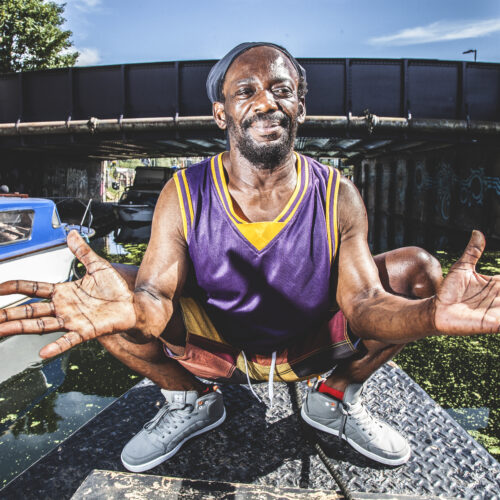 Bonjo wearing a purple and yellow basketball jersey, red shorts, and gray sneakers is squatting on a metal platform near a canal.