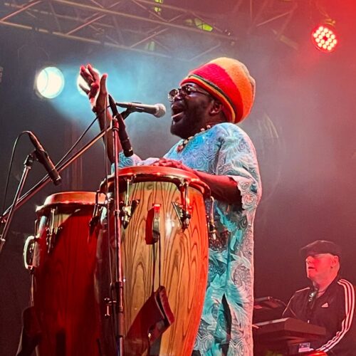 A man is standing on a stage with spotlights on him, singing into a microphone with a set of drums in front of him. There is a man sat at a keyboard behind him.