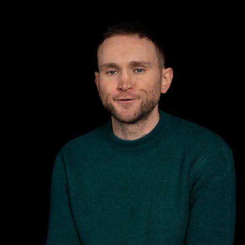 A person is shown from the torso upward, seated against a completely black background. The individual is wearing a dark teal knitted sweater with long sleeves. The lighting highlights the texture of the sweater while the rest of the scene remains simple and uncluttered.