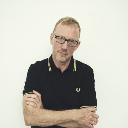 Blur's Dave Rowntree (a white male) stood with his arms crossed wearing a dark fred perry polo shirt and black framed glasses.