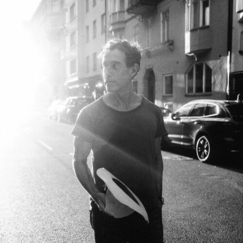 A black and white photo of Joshua Radin crossing the street. In his hand he is carrying a wide-brimmed hat, and behind him you can see apartment buildings and parked cars.