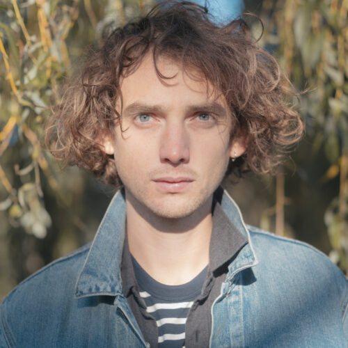 A headshot of Dan Owen with curly brown hair and a denim jacket, stood in front of some trees.