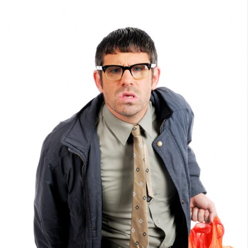 Angelos Epithemiou wearing a jacket, dress shirt, and tie, holding an orange plastic bag against a plain white background.