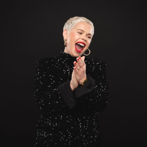 Laura Smyth wearing a black, sequined outfit and gold earrings, standing against a dark background with hands clasped together.