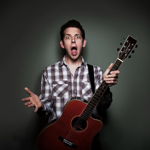 Person wearing a plaid shirt holding an acoustic guitar, standing against a dark background with one hand raised in an expressive gesture.