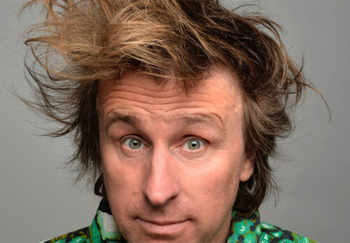 Milton Jones with wild, tousled hair wearing a brightly patterned green and black top against a plain grey background.
