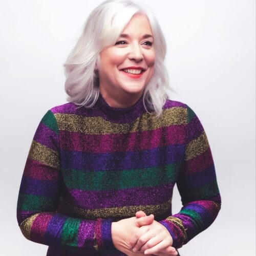 Lou Conran with shoulder‑length light hair wearing a long‑sleeved top with bold horizontal glittery stripes in purple, green, and gold, standing against a plain light background with hands loosely clasped.