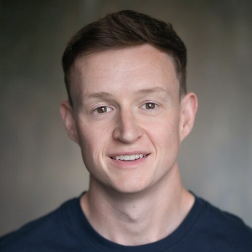 A person with short brown hair wearing a dark crew‑neck shirt, shown from the shoulders up against a softly blurred neutral background.