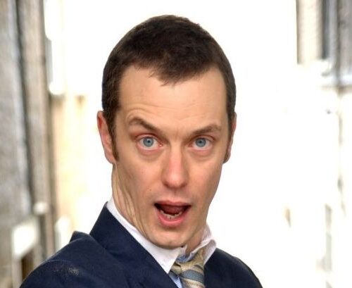 A person wearing a dark suit jacket, light shirt, and patterned tie, standing in an outdoor urban setting with tall buildings and bright light in the background.