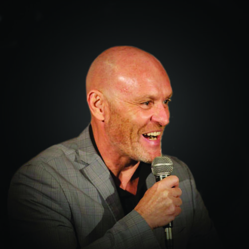 A person wearing a checked blazer and holding a microphone while speaking against a dark, softly lit background.