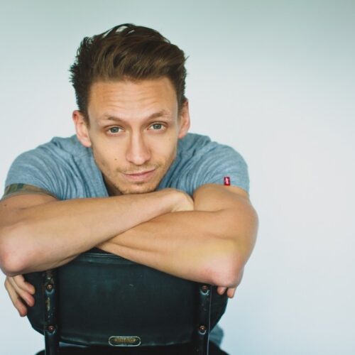 A person sitting on a backward-facing chair, resting their arms across the top of the chair back, wearing a short-sleeved grey shirt against a plain light background.