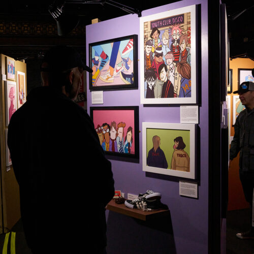 An indoor art exhibition featuring brightly colored, cartoon-style illustrations displayed on partition walls. Several visitors stand in the dimly lit space viewing the artwork, which includes framed prints, small shelves with objects, and caption cards beside each piece. The room has spotlights highlighting the art and a casual, gallery-like atmosphere.