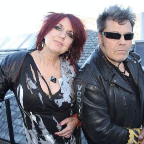 Two people stand on an outdoor rooftop area beside a railing, wearing bold patterned clothing and leather jackets, with sloped rooftops and skylights visible in the background.