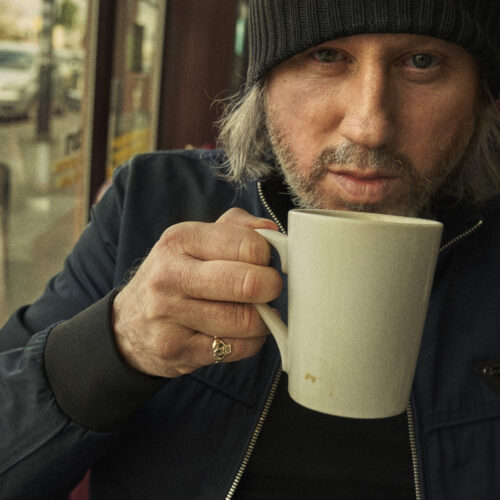 A person wearing a dark beanie and navy jacket sits indoors, holding a white ceramic mug close to their face. They wear a gold skull ring on their ring finger. Behind them is a window showing parked cars on a street and stacked chairs inside the café.