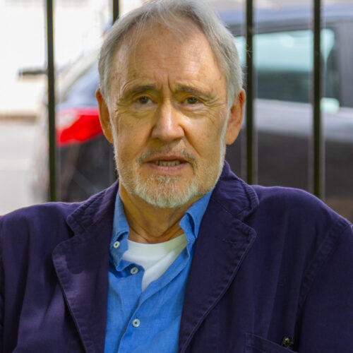 A person with short, light-colored hair is seated outdoors in front of a black metal fence. The person is wearing a blue shirt layered under a white undershirt and a dark blue jacket. A parked car and a building are visible in the background, slightly out of focus.
