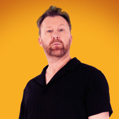 A person is shown from the torso upward against a vivid orange background. The individual is wearing a short‑sleeved black collared shirt with a textured fabric. The lighting casts soft highlights on the shirt and hair, creating a bold contrast with the smooth, warm backdrop.