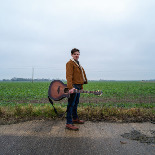 A person stands on a rural roadside holding an acoustic guitar by the neck, with a green field stretching out behind them under an overcast sky. The person is wearing a tan jacket, dark trousers and boots. Power lines run horizontally across the landscape, and the ground in the foreground appears wet and textured.