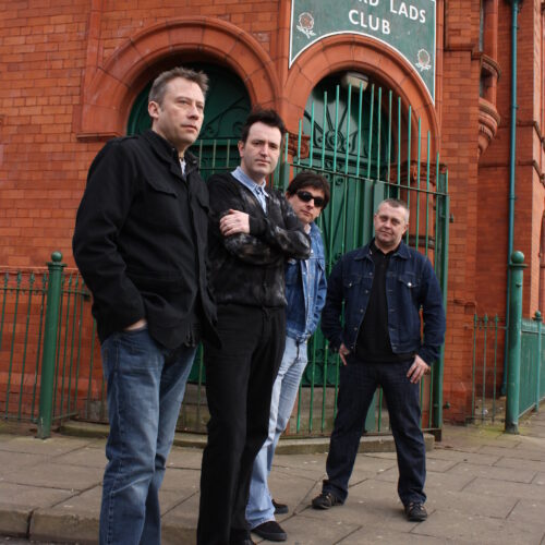Four men stand on a pavement in front of the Salford Lads Club, a historic red‑brick building with green iron gates and a large sign above the entrance. The men pose casually, wearing jackets and jeans, with the building’s distinctive architecture and fencing visible behind them.