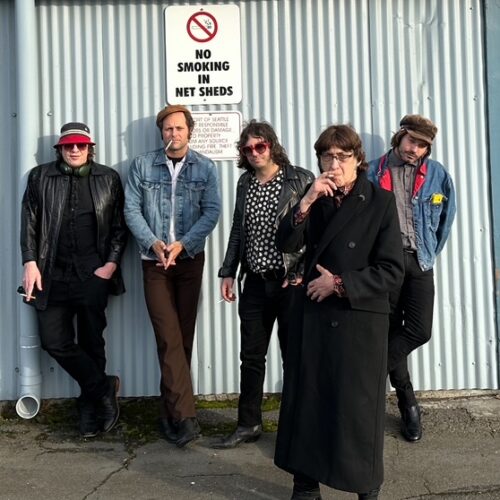 Five men stand casually against a corrugated metal wall beneath a “No Smoking in Net Sheds” sign. They wear a mix of leather jackets, denim, hats, and sunglasses. One man in a long black coat stands slightly forward, smoking, while the others lean or stand relaxed, each with a distinct rock‑and‑roll style.