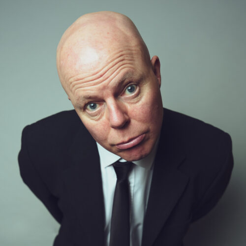 Studio portrait of a person leaning slightly forward toward the camera against a light grey background, wearing a dark suit jacket, white shirt, and black tie, lit evenly from the front.