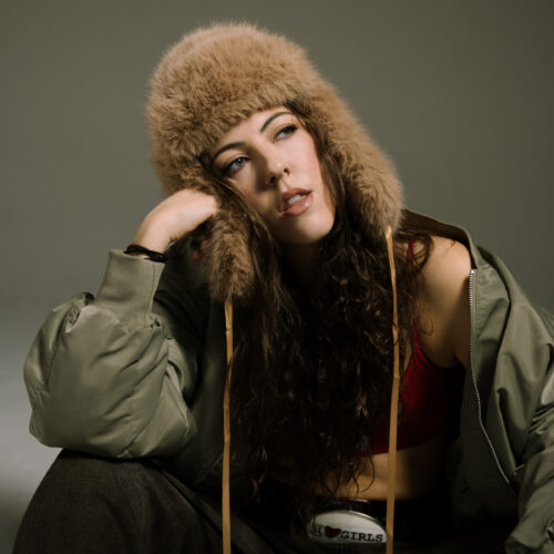 Studio portrait of a person seated against a plain background, wearing a fur-lined hat and oversized jacket, with long hair and one hand resting against the side of the face.