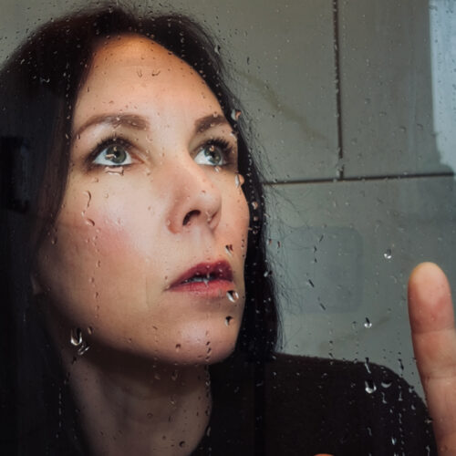 Close-up of a person’s face seen through rain-speckled glass. Water droplets cling to the surface, slightly blurring the features. The person looks upward with one hand raised near the glass, with a neutral indoor background visible behind them.