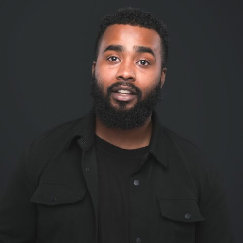 A person with short curly hair and a full beard faces the camera in a studio portrait. They wear a black shirt and black jacket against a dark grey background, with even lighting highlighting their facial features.