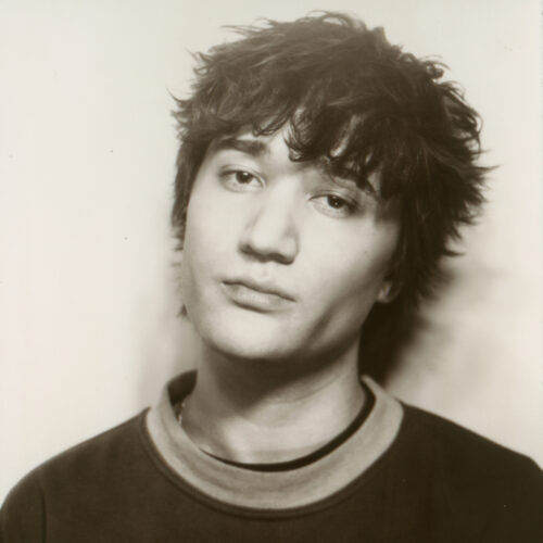 Soft-focus black-and-white portrait showing a person with short, textured hair, looking toward the camera, wearing a dark top with faint text visible, set against a minimal light background.