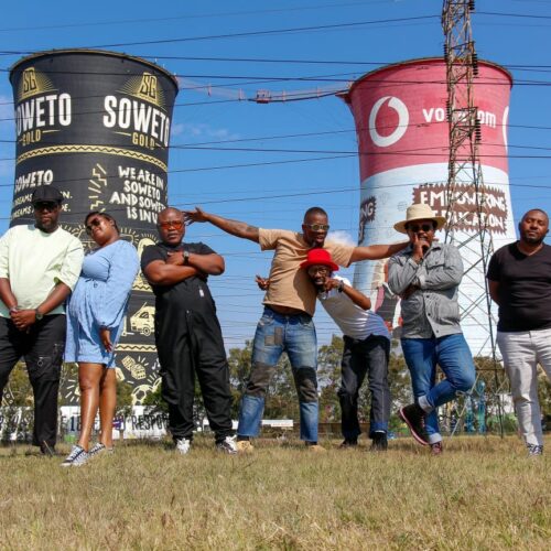 Seven members of BCUC standing in a loose line on grassy ground, posing in front of two large painted cooling towers with visible text and graphics, under a bright blue sky with power lines crossing above.