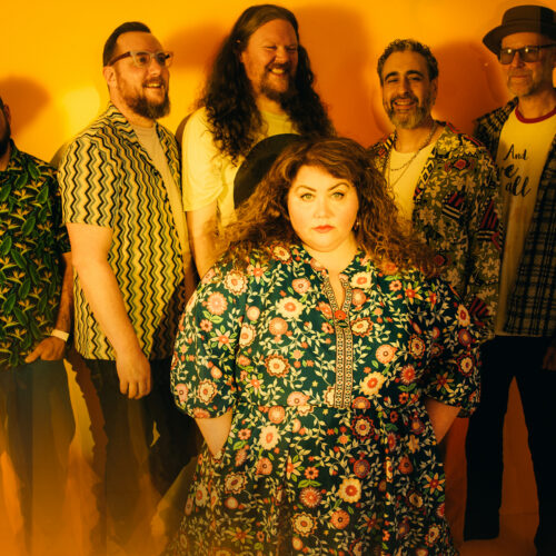 Group portrait of a band standing against a warm yellow-orange background, with one person in the foreground wearing a floral dress and five others positioned behind in patterned shirts and jackets.