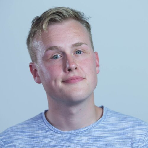 Studio portrait showing a person with short light-coloured hair wearing a light blue striped knit top, photographed against a plain pale blue background.