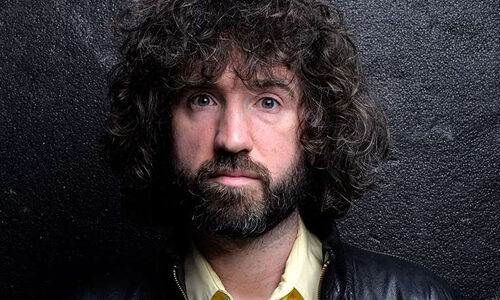 Close-up promotional photograph of a performer with curly hair and a beard, wearing a dark jacket over a light-coloured shirt. The image is set against a textured black background and is tightly cropped around the head and shoulders.