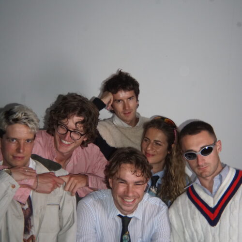 Group portrait of six people posed closely together against a plain white wall. One person sits at the front wearing a light blue striped shirt and patterned tie. Five others stand or lean behind and beside the seated person. Clothing includes a pink button-down shirt, a cream-colored jacket, a white sweater with red and blue trim, and a light sweater. Two people wear glasses, including one with dark round frames and one with white-framed sunglasses. The photo is lit with direct flash, creating strong shadows on the wall behind the group.