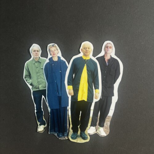 Promotional group photograph showing four band members standing close together against a plain black backdrop. The group wear contrasting outfits including a green jacket, a long blue coat, a dark tunic with a bright yellow shirt, and a black shirt with sunglasses.