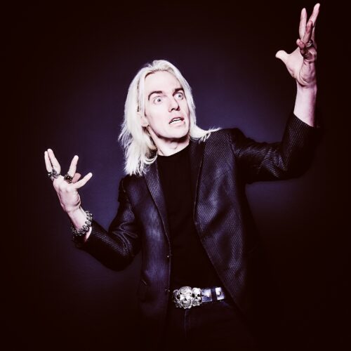 Studio portrait of a person with long light-coloured hair wearing a dark jacket, gesturing with both hands and wearing rings and bracelets against a dark background.