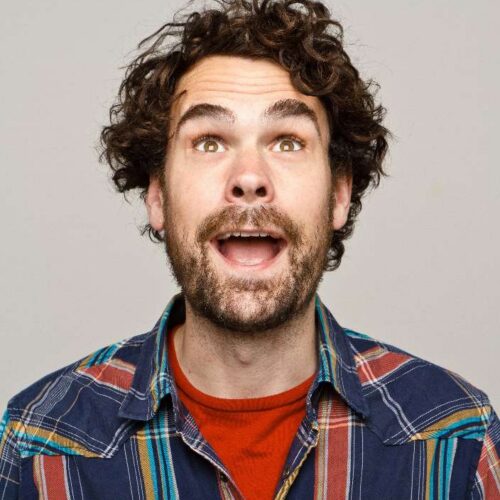 Head-and-shoulders portrait of a person with curly hair and a beard wearing a blue checked shirt and red top, photographed against a light grey background.