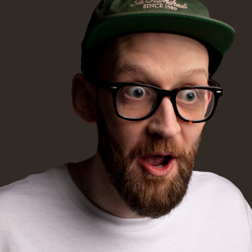 Head-and-shoulders portrait of a person wearing black-framed glasses, a green cap and a white T-shirt, photographed against a dark background.